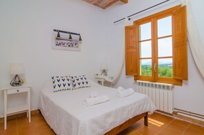 A bedroom with a bed, two bedside tables, a desk, a window with curtains, a radiator, and a wall-mounted lamp.