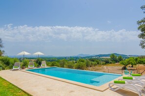 A swimming pool with lounge chairs and umbrellas, surrounded by greenery and hills in the background.
