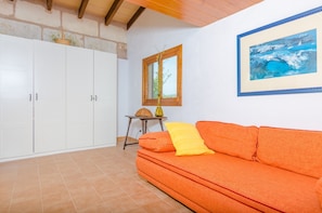 Een kamer met een betegelde vloer, een witte garderobe, een oranje bank met een geel kussen, een klein houten tafeltje met een vaas, en een lijst met een afbeelding aan de muur.