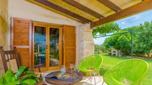 Un porche cubierto con muebles de madera, una mesa con una revista y vasos, y vistas a un jardín y árboles.