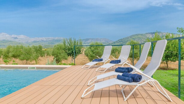 Una terraza de madera con cuatro sillas reclinables blancas, cada una con una toalla azul, que da vistas a una piscina y un paisaje montañoso.