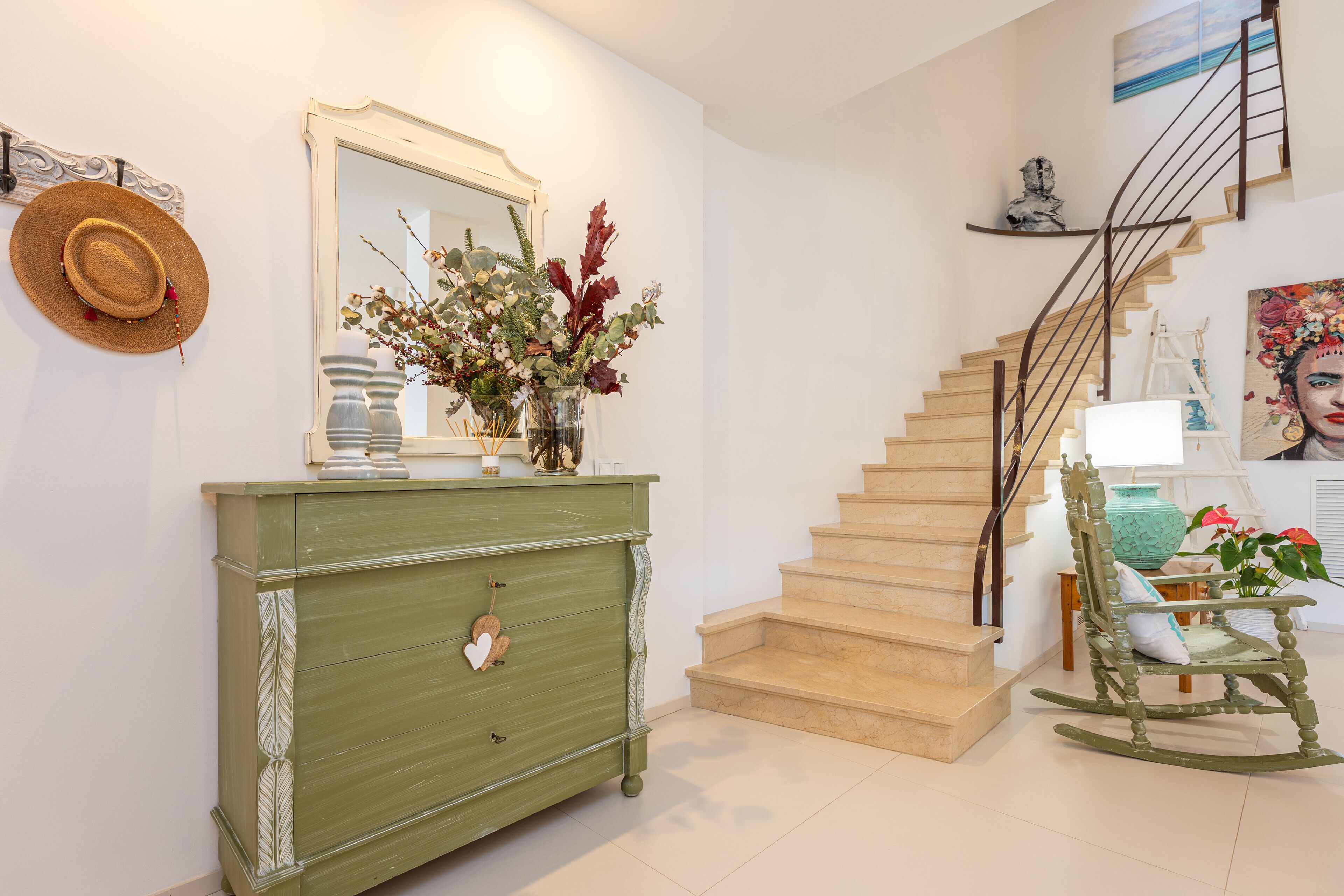 A staircase with a wooden finish, a green dresser with decorative elements, a rocking chair, and a mirror with a floral arrangement.