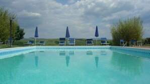 Une piscine à l’eau d’un bleu cristallin, entourée de chaises longues et de parasols, avec en toile de fond un ciel nuageux et des collines verdoyantes.