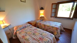 Une chambre avec deux lits, chacun agrémenté d’une literie à motifs floraux, une table de chevet, une lampe et une fenêtre donnant sur un paysage verdoyant.