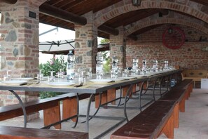 Una larga mesa de comedor dispuesta con platos, vasos y cubiertos, rodeada de bancos en un espacio al aire libre con paredes de piedra.