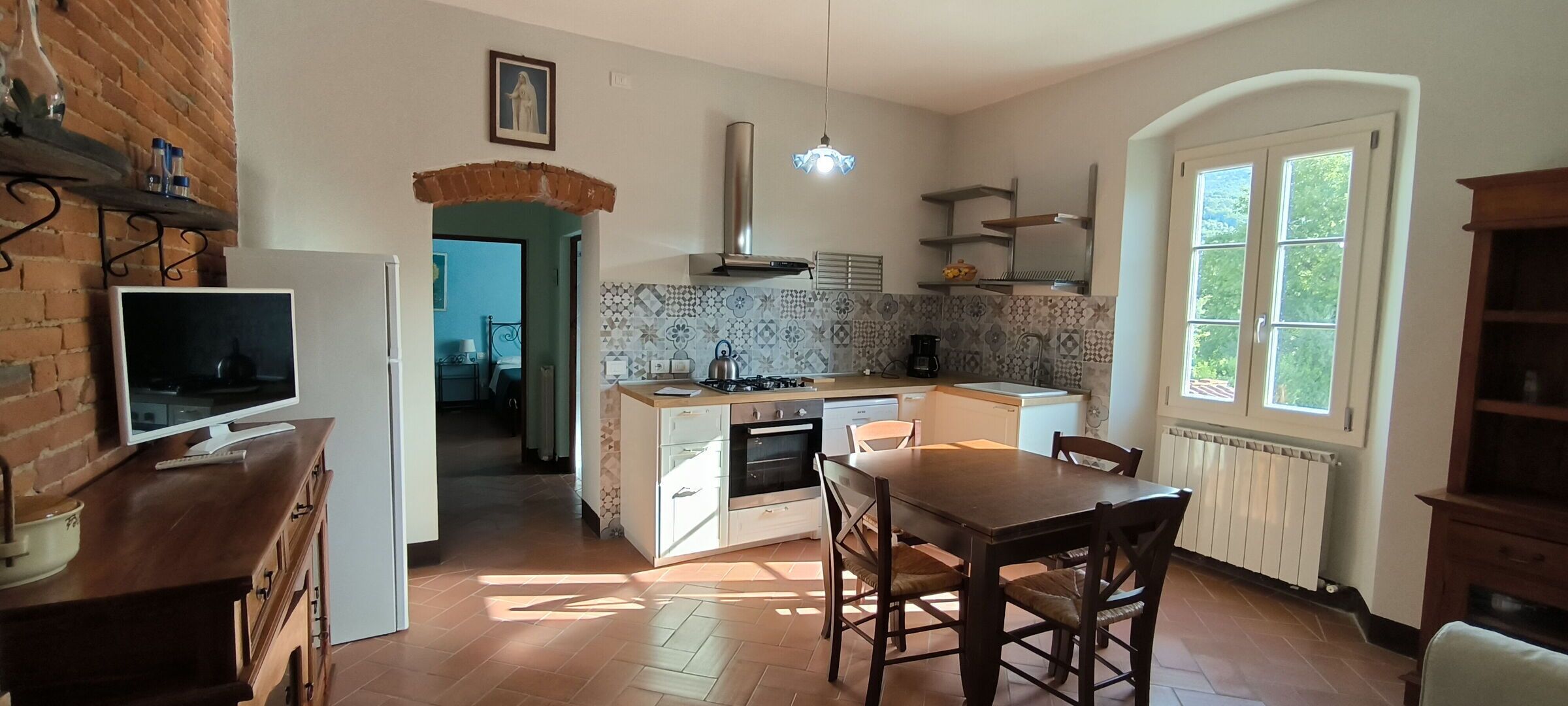 A kitchen with a TV on a wooden counter, a dining table with chairs, and a window with a view of greenery.