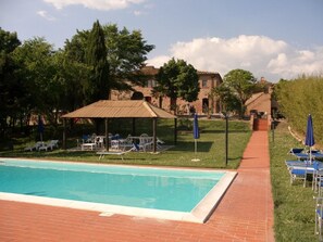 A swimming pool with lounge chairs and a gazebo, surrounded by greenery and a brick building in the background.