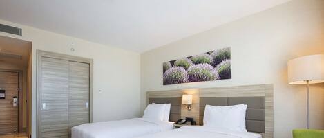 Une chambre d’hôtel avec deux lits, un fauteuil vert, une table, un tableau accroché au mur et une porte.