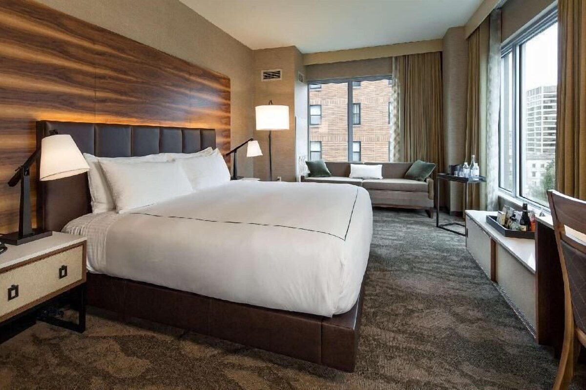 Habitación de hotel con una cama grande, un sofá, un escritorio con lámpara y vistas a un edificio de ladrillo.