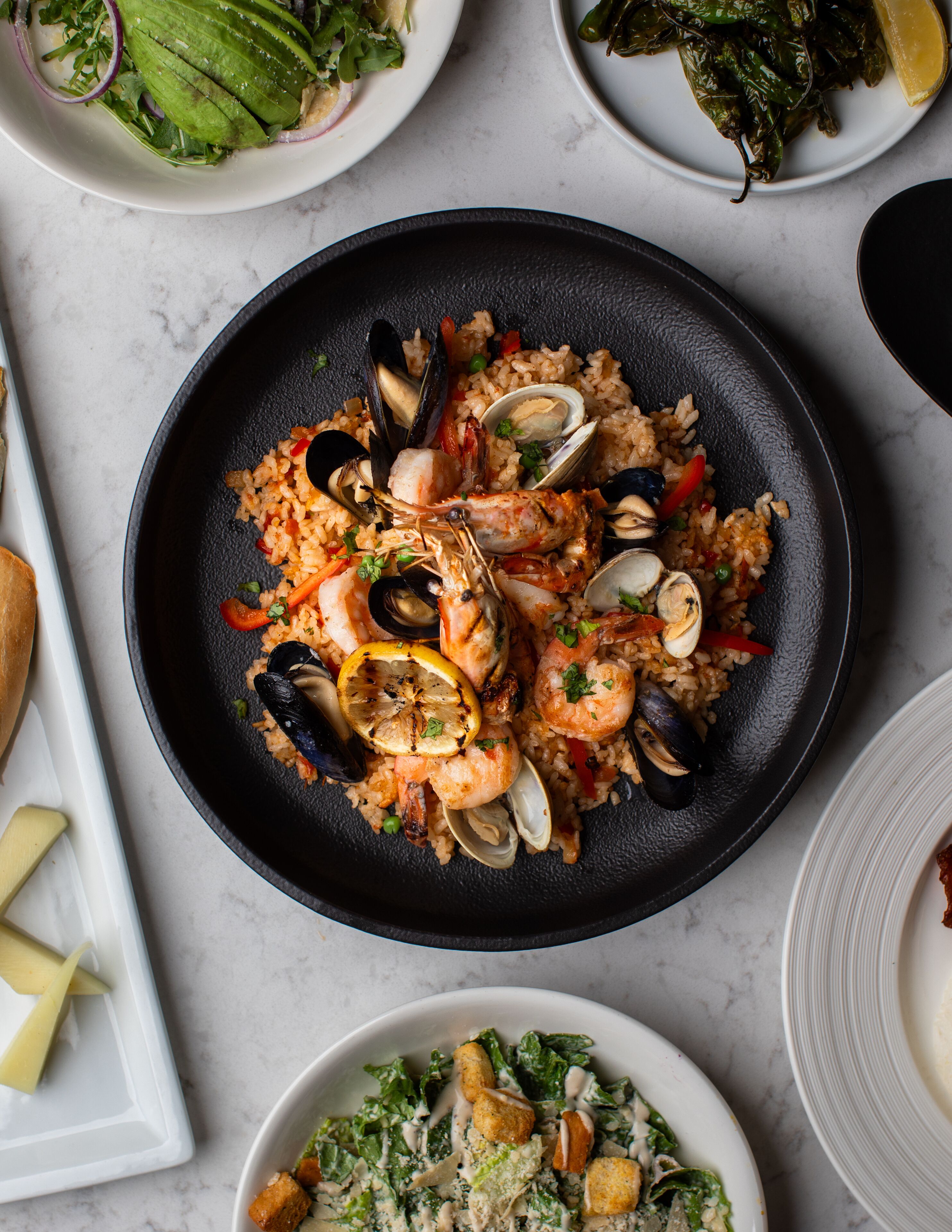 A seafood paella dish with mussels and shrimp on a black plate, surrounded by other dishes.
