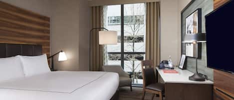 Ein Hotelzimmer mit Bett, Schreibtisch, Stuhl, Lampe und einem großen Fenster mit Vorhängen.