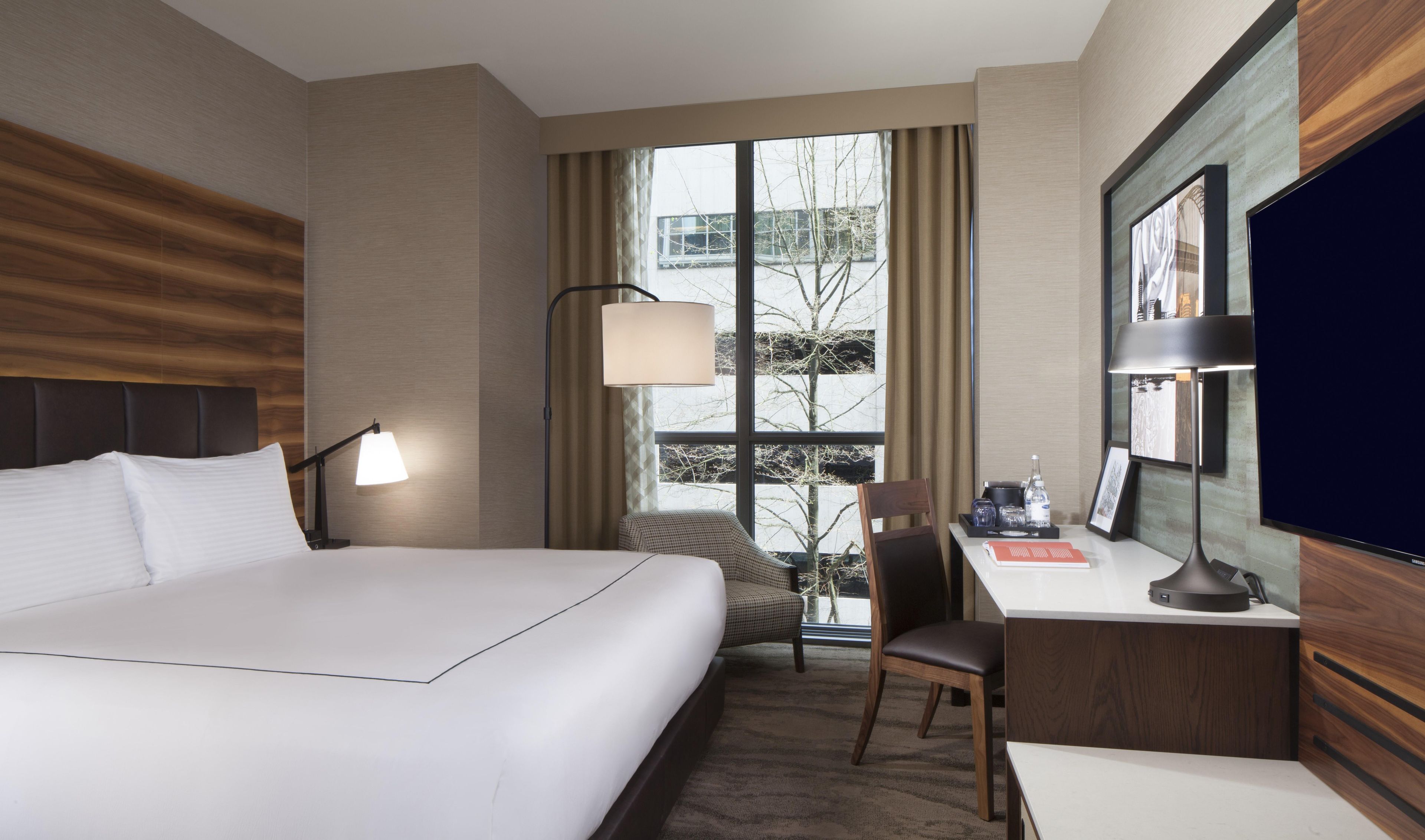 Quarto de hotel com uma cama grande, uma escrivaninha com cadeira, uma luminária, televisão e uma janela com cortinas.