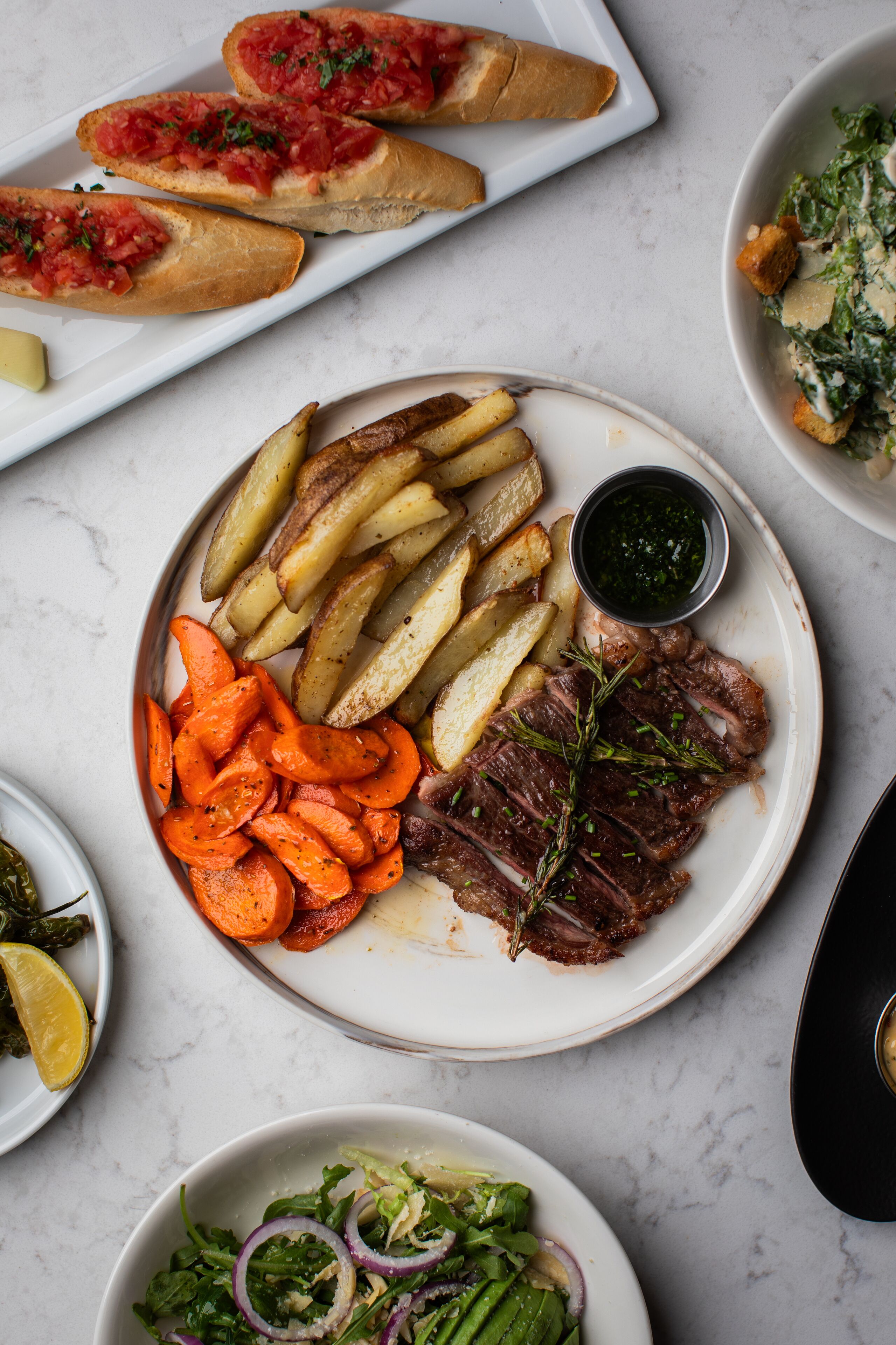 Une assiette contenant un steak, des pommes de terre rôties et des carottes, accompagnée d’un bol de salade et d’une petite portion de sauce.