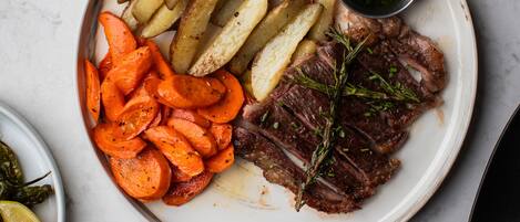 Une assiette contenant un steak, des pommes de terre rôties et des carottes, accompagnée d’un bol de salade et d’une petite portion de sauce.