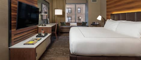 A hotel room with a large bed, a desk with a flat-screen TV, a seating area with a sofa and a chair, and a window with a view of a brick building.