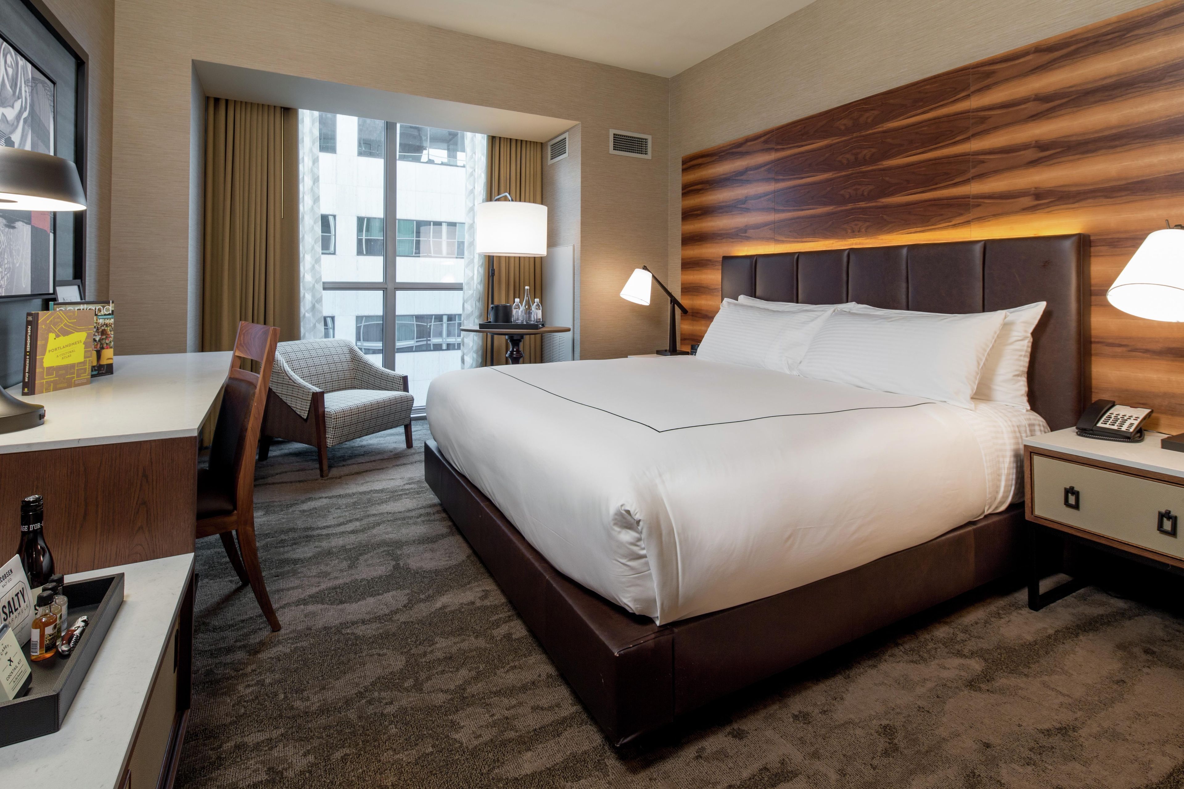 A hotel room with a large bed, bedside lamps, a desk with a chair, and a window with curtains.