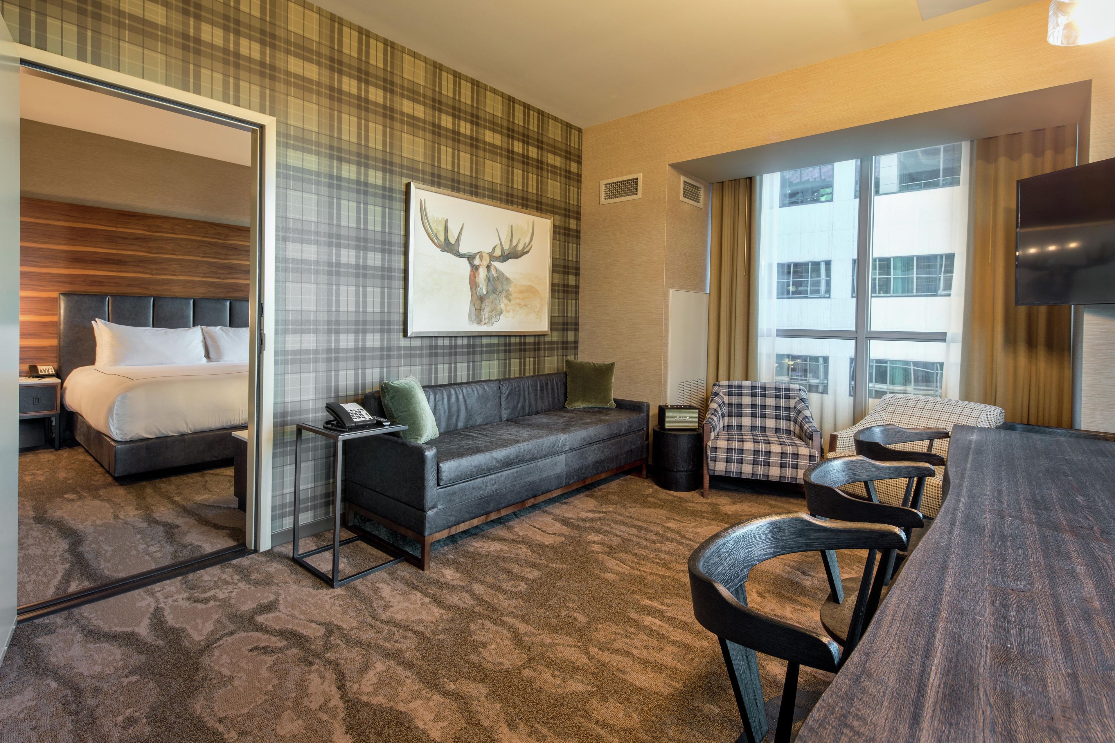 A hotel room with a bed, a sofa, a dining table with chairs, a television, and a large window.