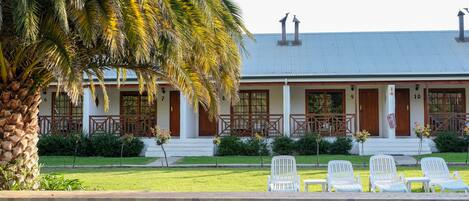 Zona de piscina con una palmera, un edificio con puertas numeradas y sillas de descanso blancas.