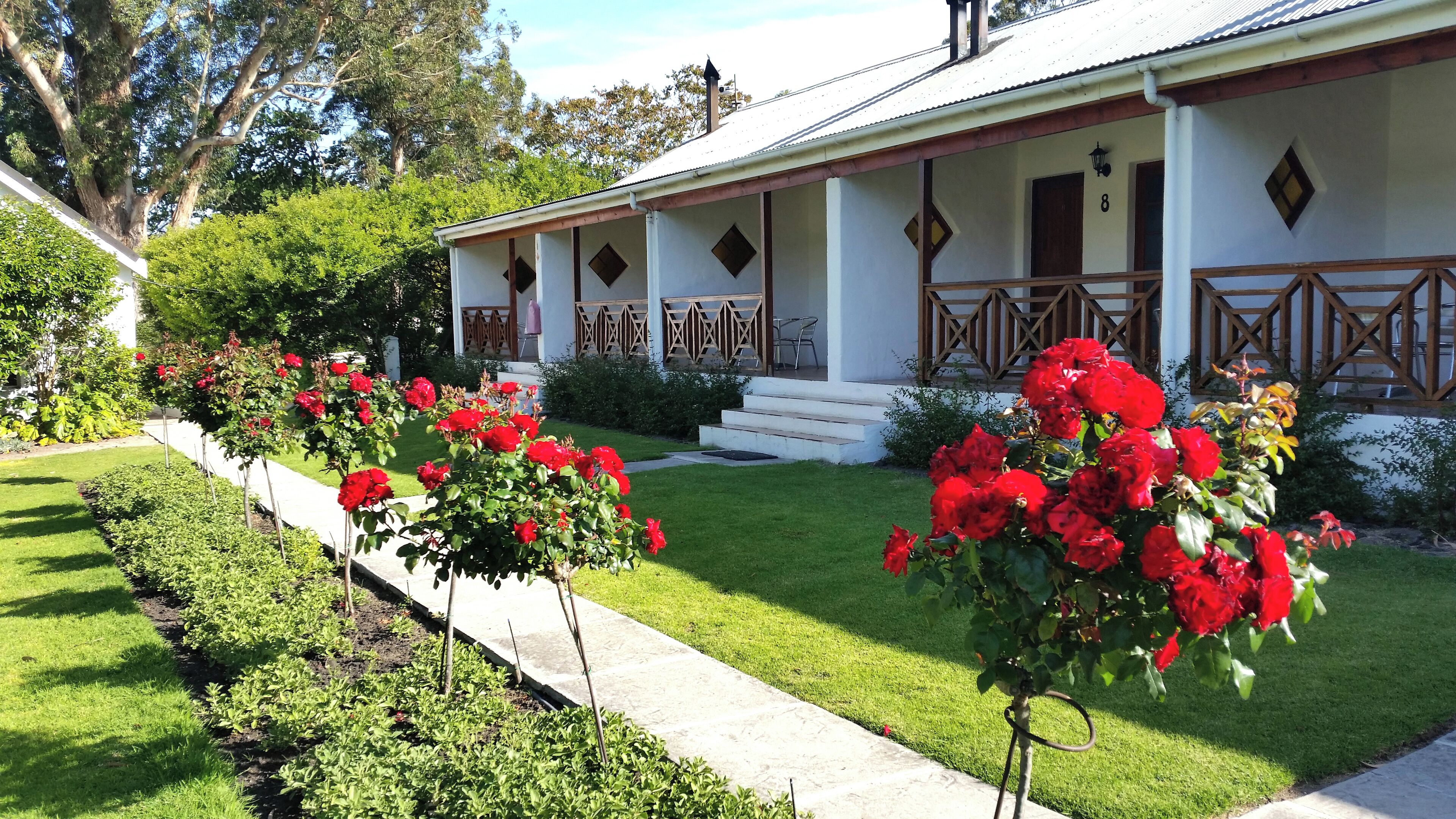 Una casa con fachada blanca, un porche con barandilla de madera y un jardín bien cuidado con flores rojas.