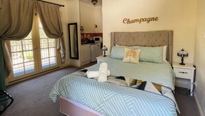 Ein Schlafzimmer mit Bett, Nachttisch, Deckenventilator, Fenster mit Vorhängen und einer Küche im Hintergrund.