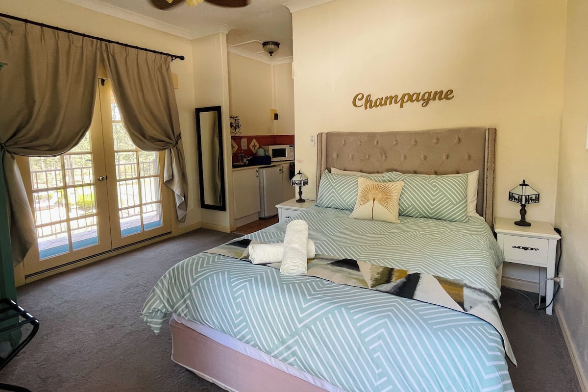 A bedroom with a bed, a nightstand, a ceiling fan, a window with curtains, and a kitchen area in the background.