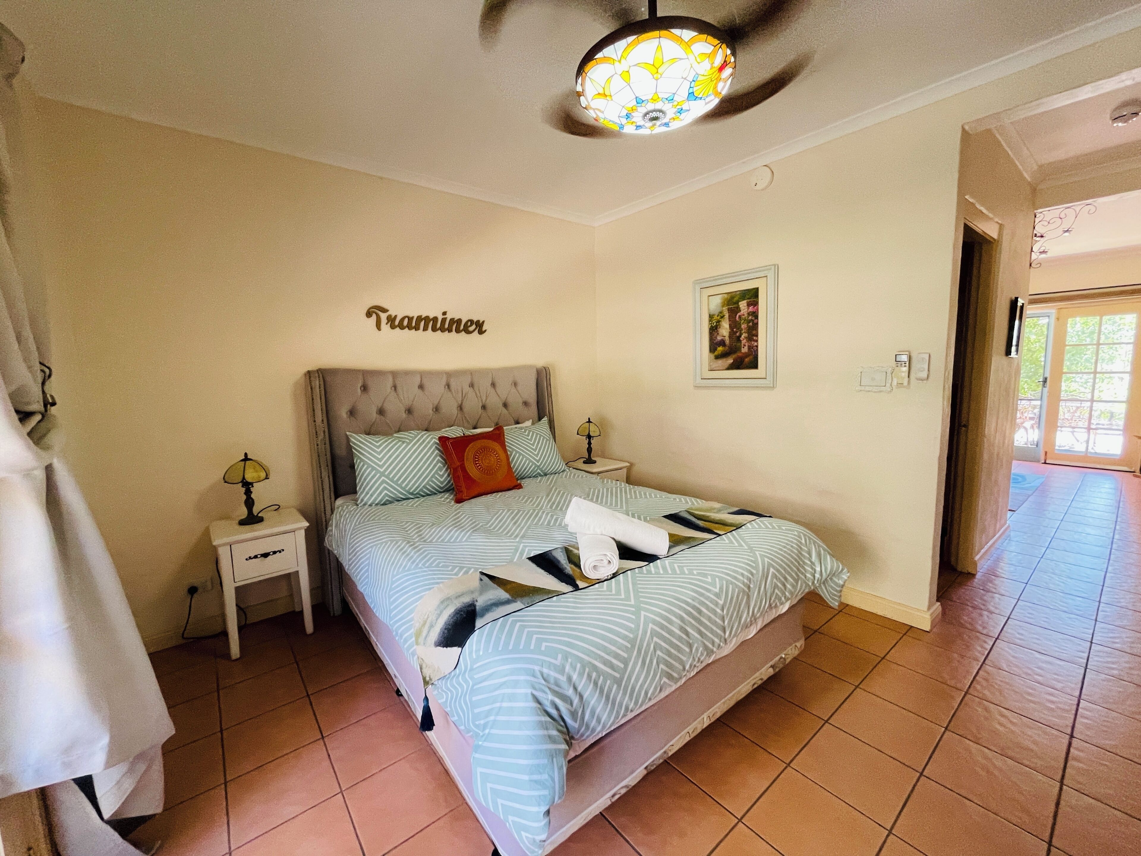 Un dormitorio con cama, mesita de noche, lámpara, ventilador de techo, un cuadro enmarcado y una puerta que da a otra habitación.