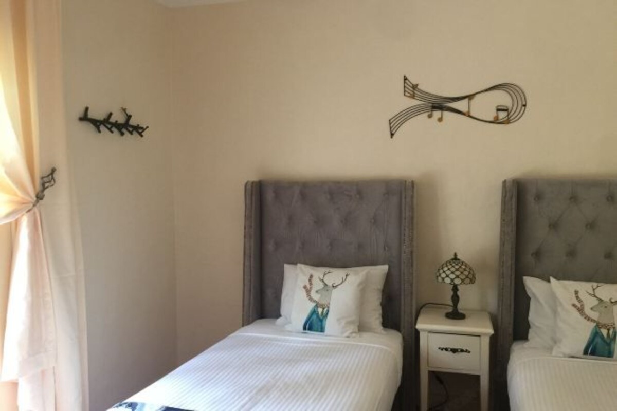 A hotel room with two beds, each with a grey headboard and white bedding. There is a small bedside table with a lamp and a decorative wall piece above the beds.