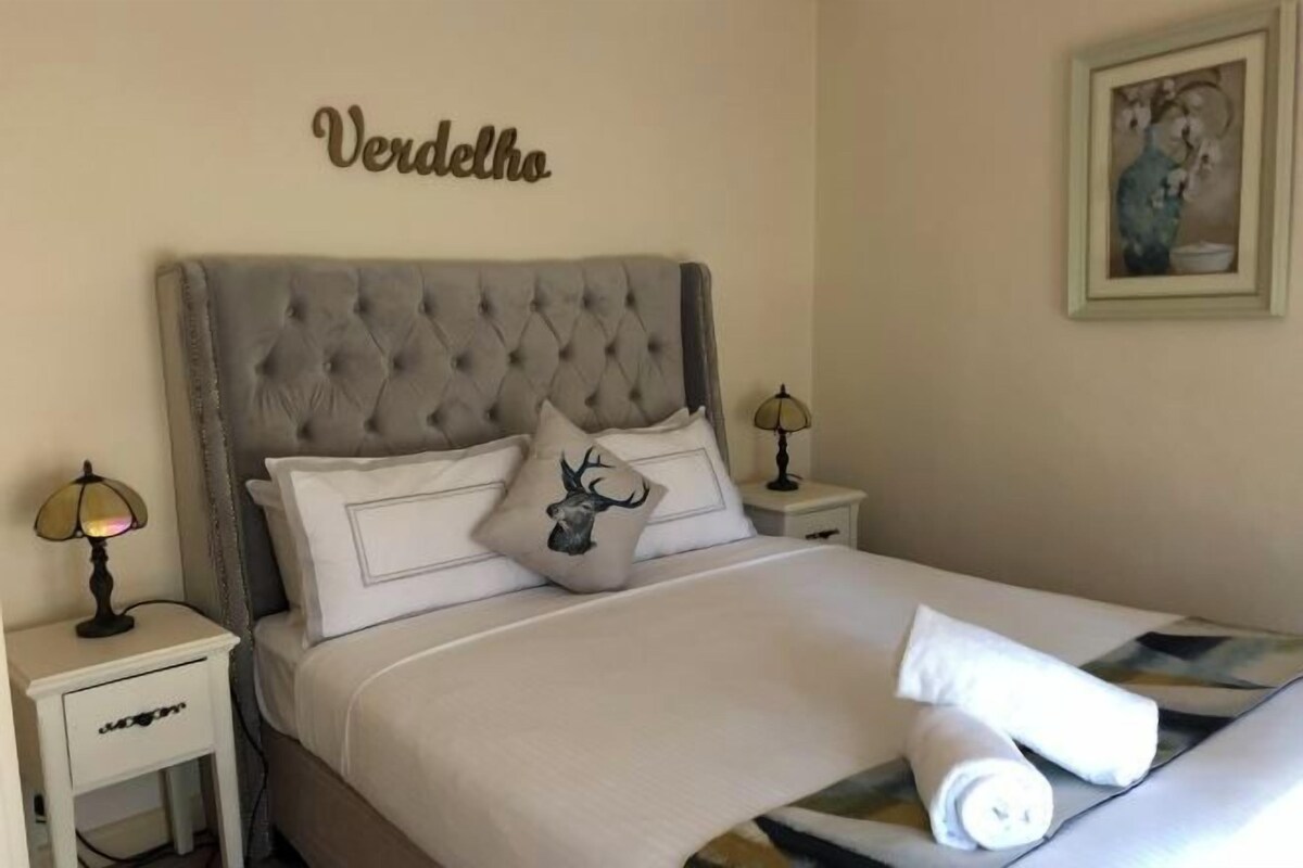 A neatly made bed with a grey tufted headboard, white bedding, and a decorative pillow with a deer head. There are two bedside tables with lamps, a framed artwork on the wall, and a sign above the bed.