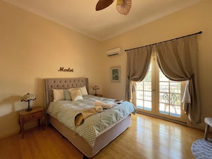 A bedroom with a tufted headboard, a bed with striped bedding, a bedside table with a lamp, a window with curtains, and a wall-mounted air conditioner. - Villa Provence (Pokolbin)