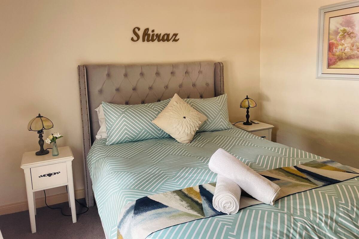 A neatly made bed with patterned bedding, a headboard, two bedside tables with lamps, and a wall-mounted sign that reads "Shiraz".