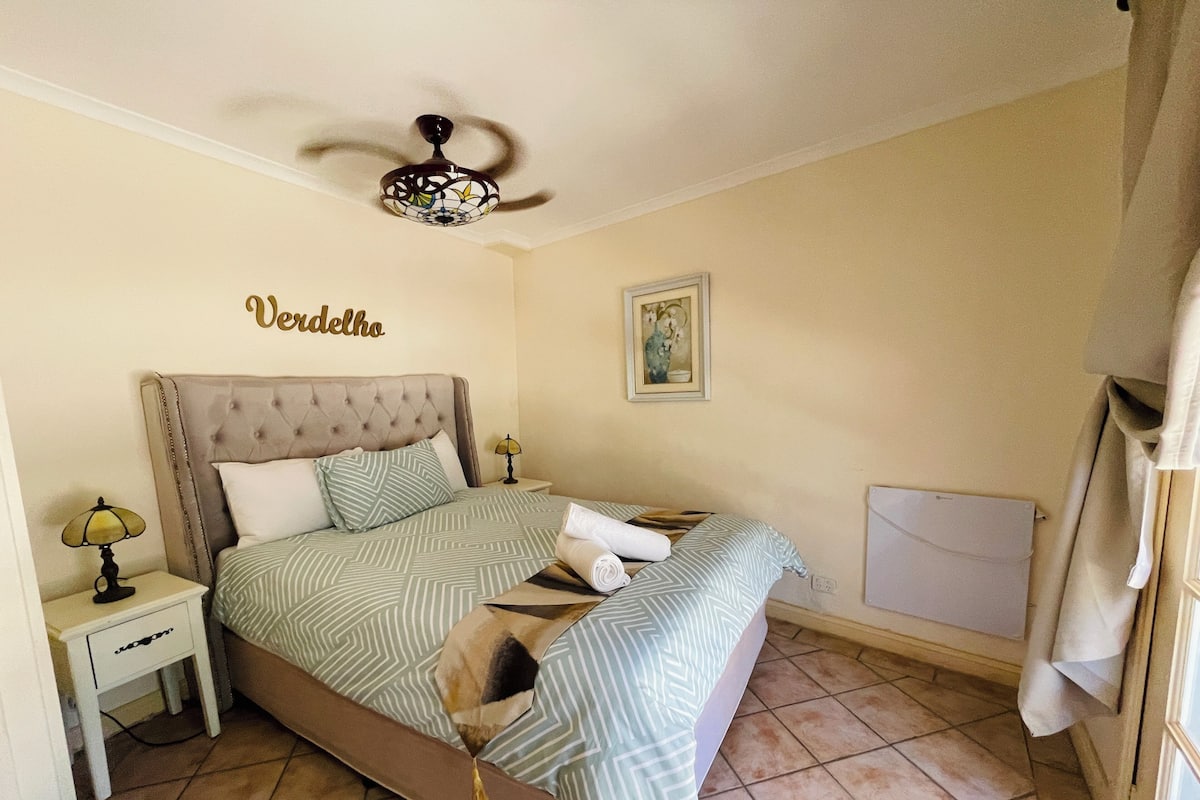 A bedroom with a bed, a nightstand, a ceiling fan, and a wall-mounted artwork.