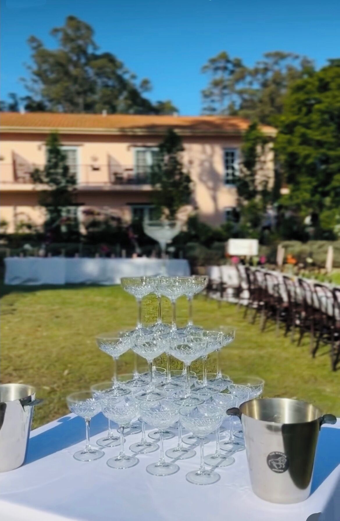 A table set with crystal glasses and silver pitchers, outdoors with a building and trees in the background.