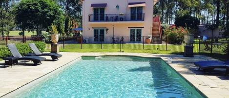 Una casa de dos pisos con piscina, rodeada por una valla y árboles.