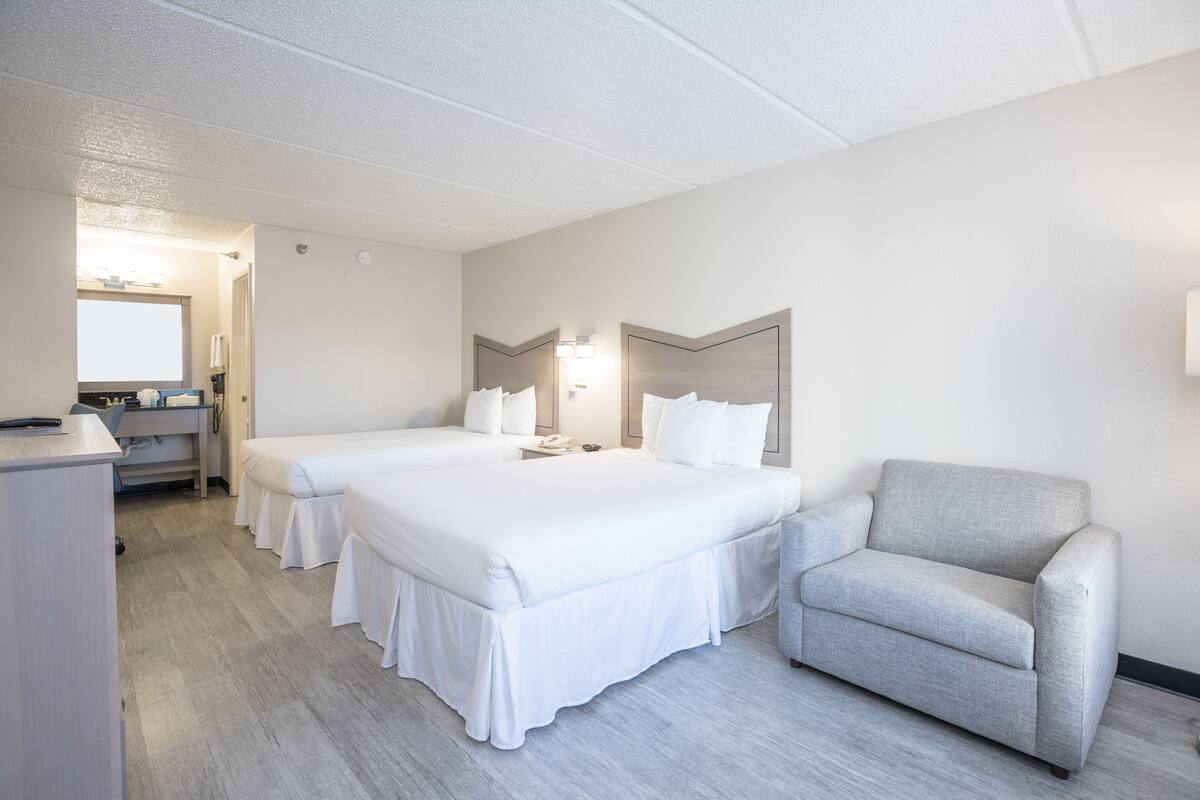 Ein Hotelzimmer mit zwei Betten, einem Schreibtisch, einem Stuhl und einem durchgehenden Türrahmen einsehbarer Badezimmer.