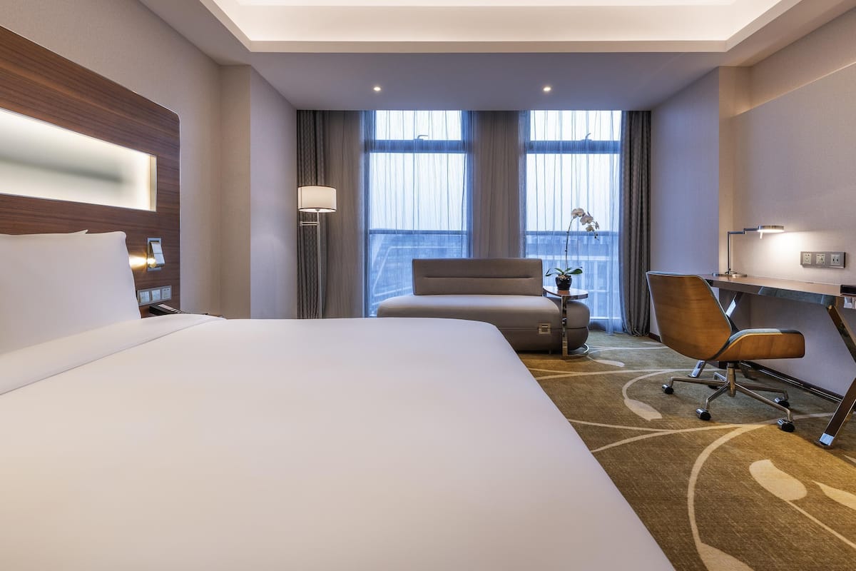 Ein Hotelzimmer mit einem großen Bett, einem Schreibtisch mit Stuhl, einer Lampe und Blick auf die Stadt durch große Fenster.