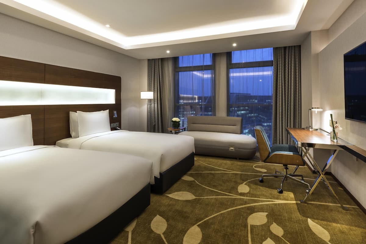 Ein Hotelzimmer mit zwei Betten, einem Schreibtisch, einem Stuhl, einer Lampe und Blick auf die beleuchtete Stadt.