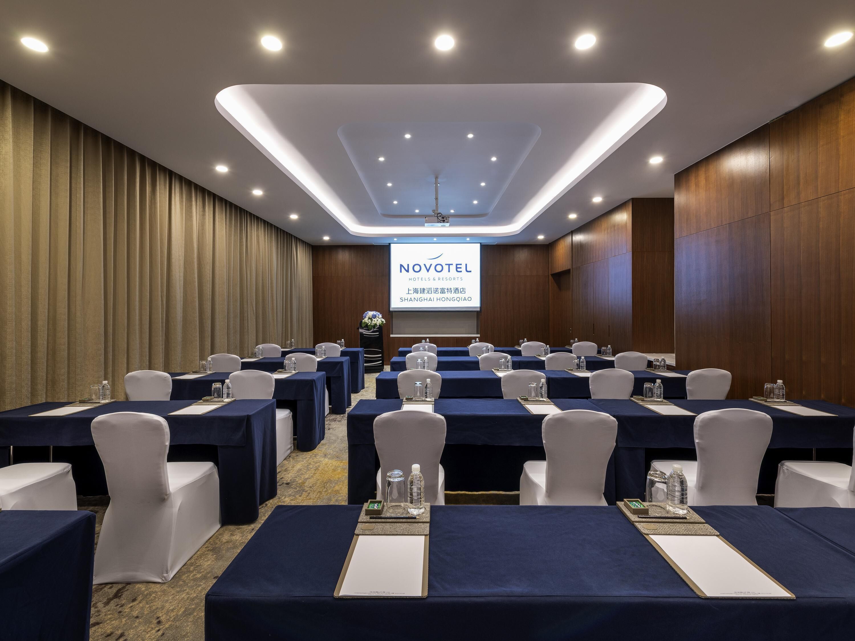 A conference room with tables, chairs, and a projector screen displaying 'NOVOTEL HOTEL & RESORTS'.