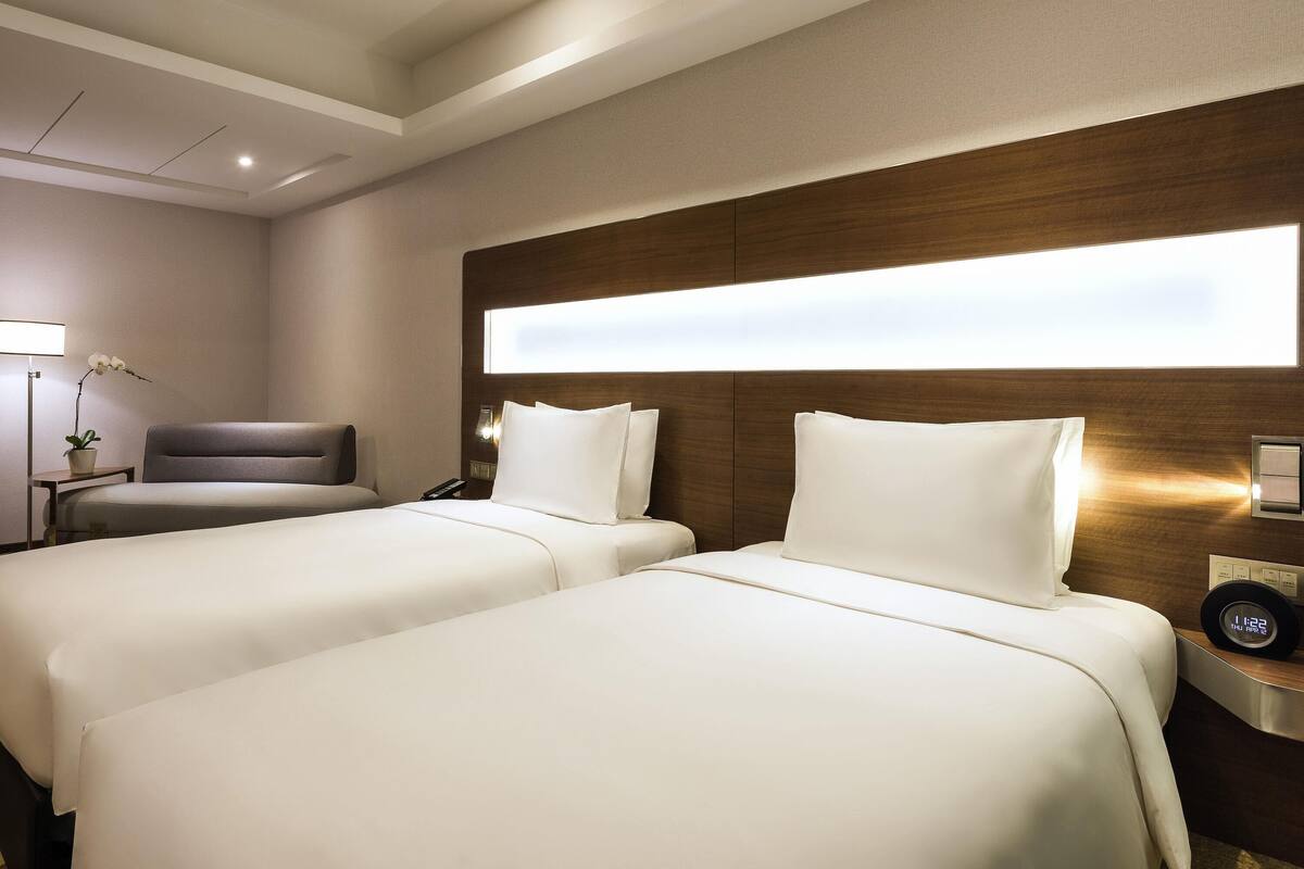 Une chambre d’hôtel avec deux lits, un bureau, une chaise, une table de chevet avec une horloge et une lampe fixée au mur.