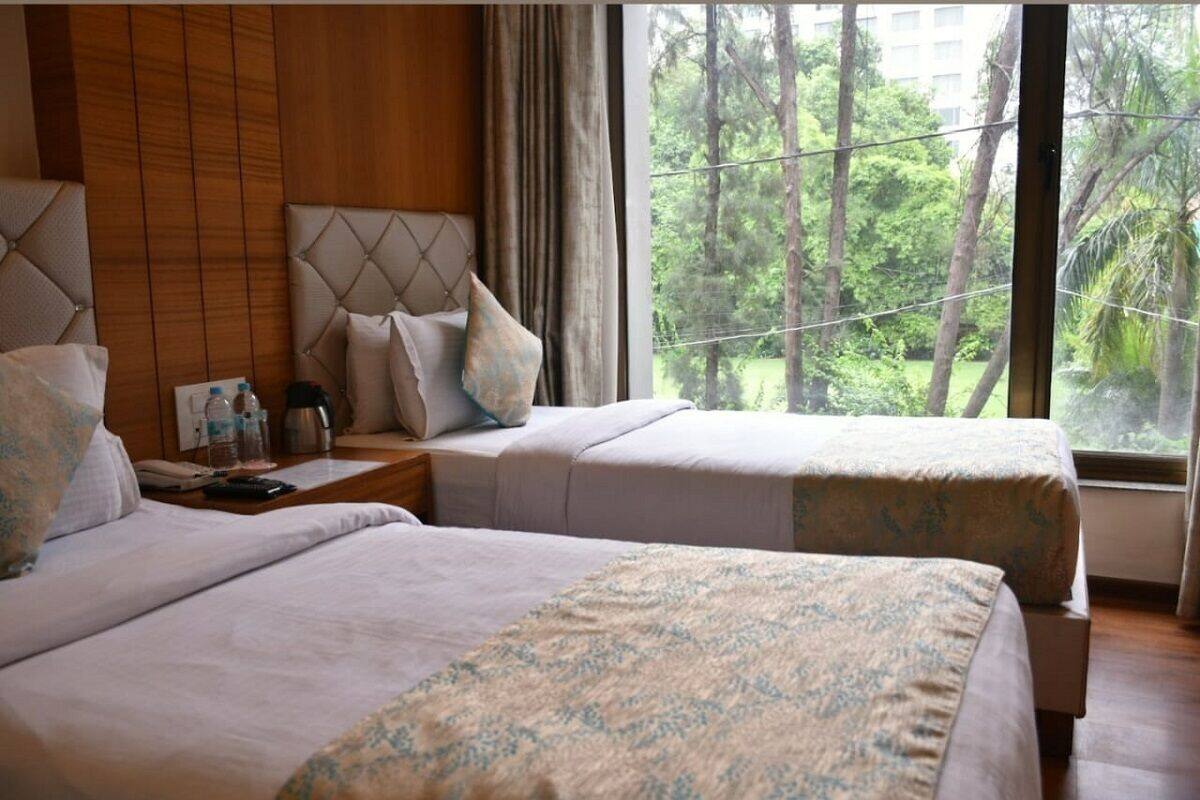 A hotel room with two beds, a wooden headboard, a nightstand with a coffee maker, and a view of trees through large windows.