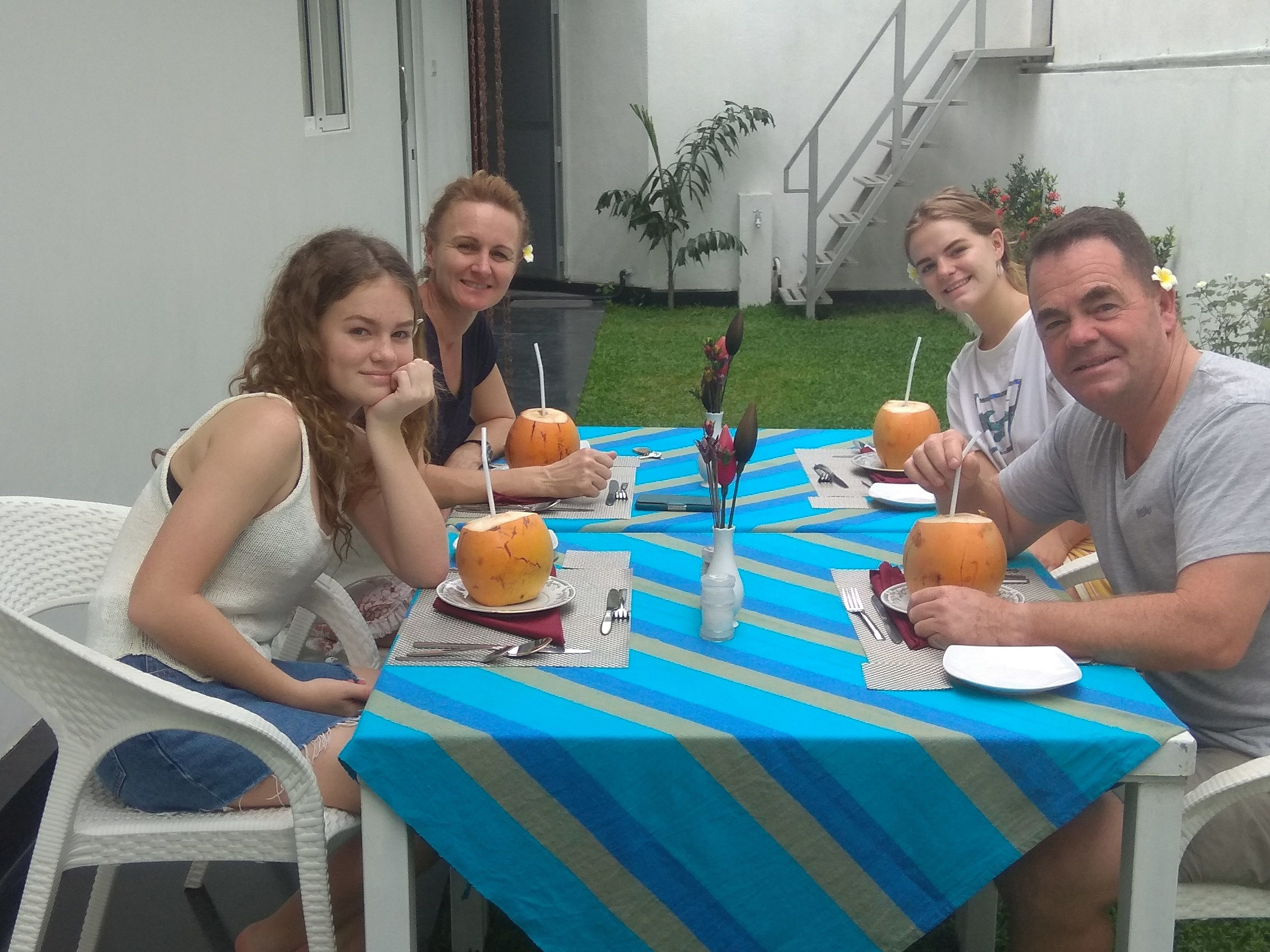 A group of people seated at an outdoor table with a blue-striped tablecloth, enjoying drinks and a meal.