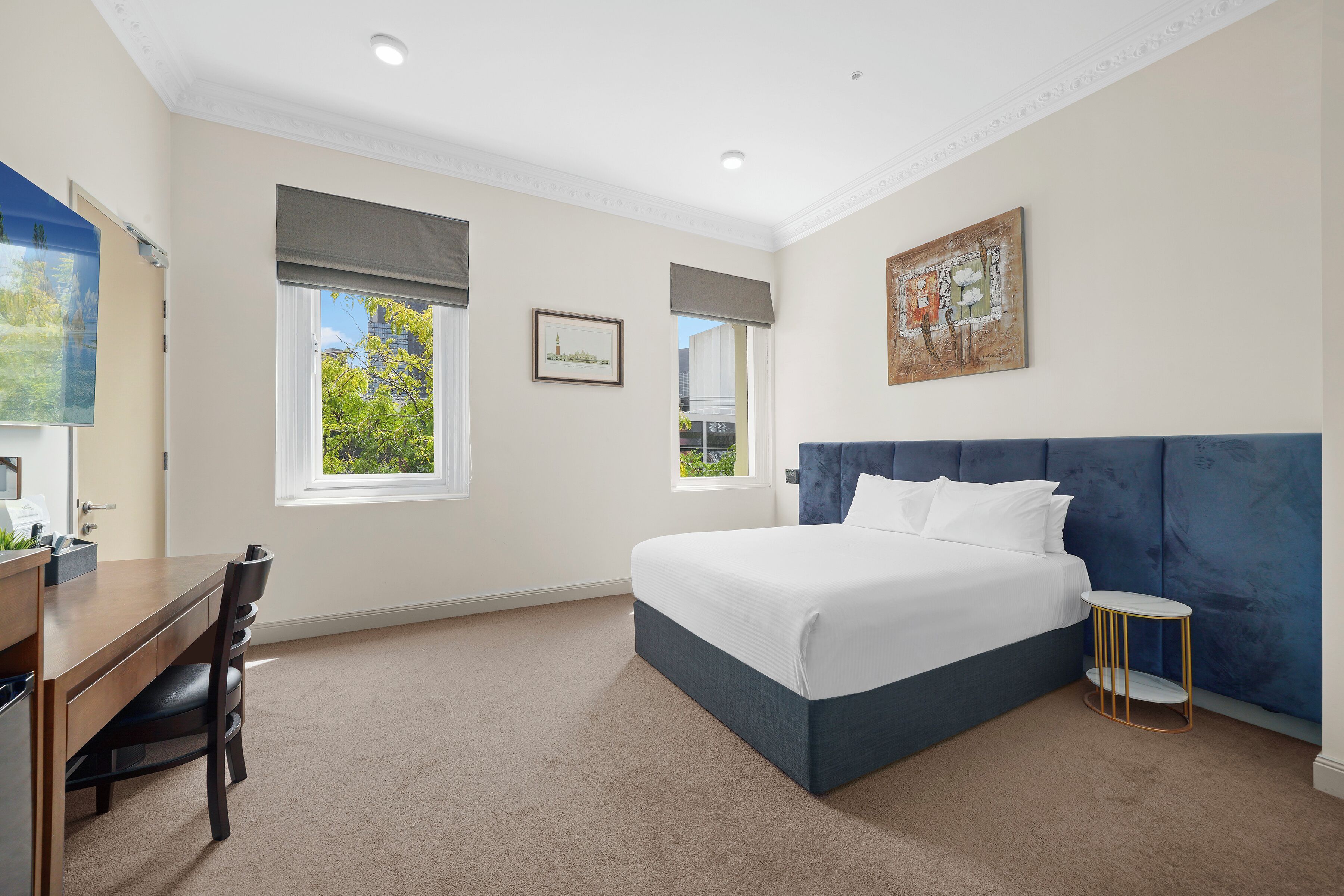 Ein Schlafzimmer mit Bett, Schreibtisch und Stuhl. Zwei Fenster mit Jalousien, ein Bild an der Wand und ein kleiner runder Tisch mit Stuhl sind ebenfalls vorhanden.