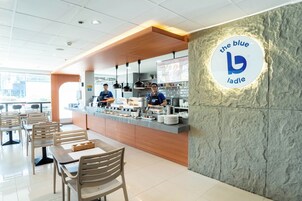 Interior de una cafetería con mostrador, zona de mesas y una pared con un logotipo.