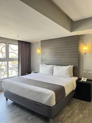 Quarto de hotel moderno com uma cama grande, mesa de cabeceira com telefone e vista da cidade pela janela.