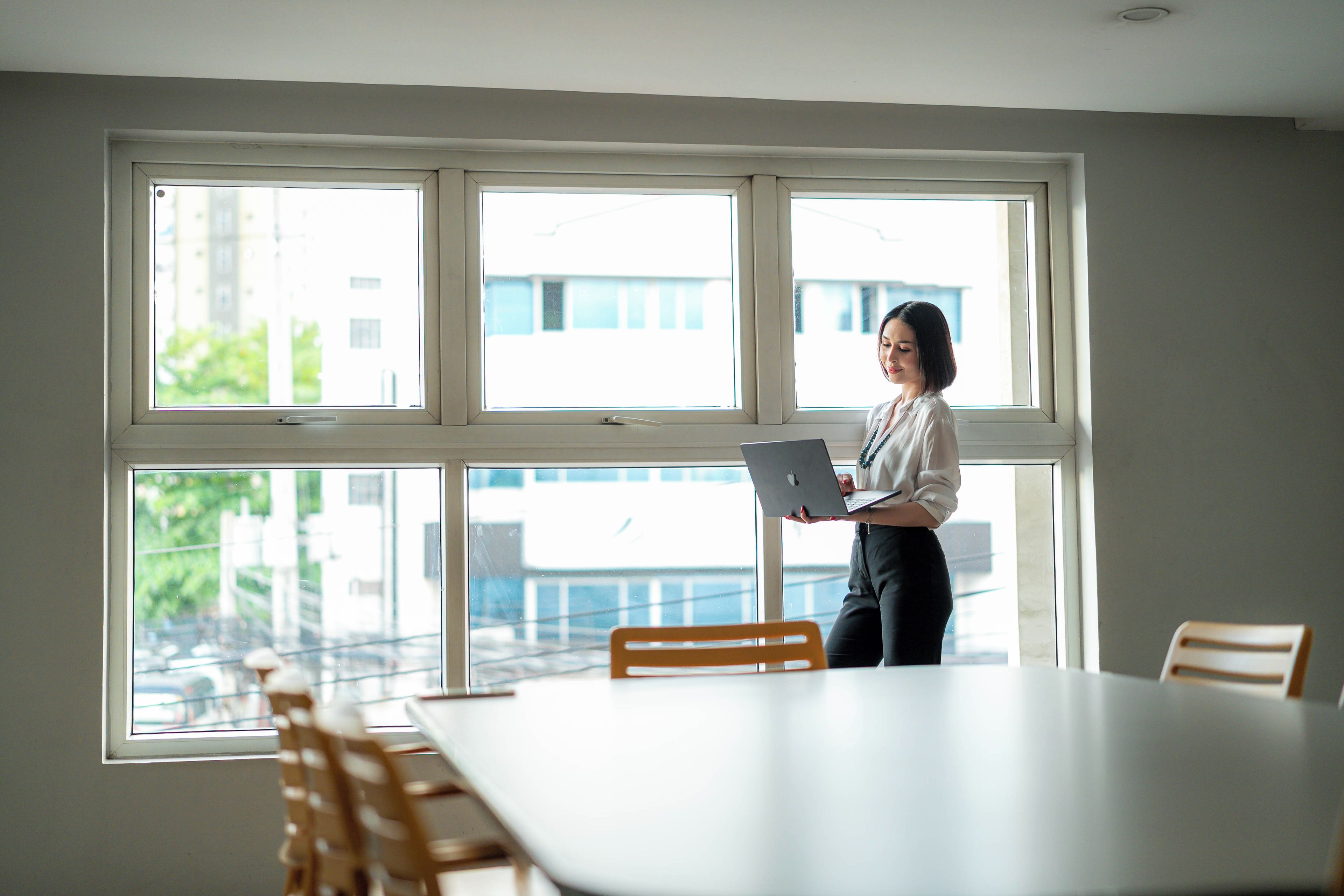 A person standing by a window with a laptop, in a room with a table and chairs.