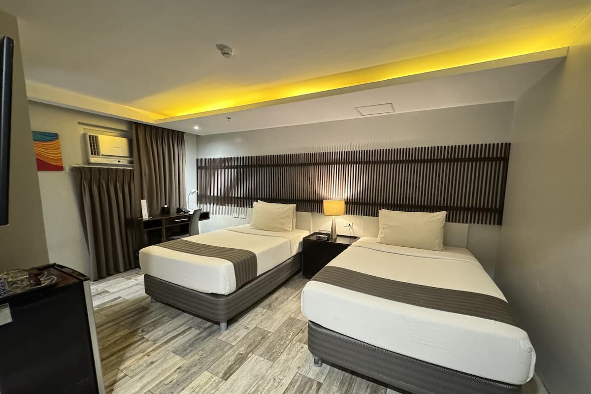 Quarto de hotel com duas camas, uma escrivaninha, uma cadeira, uma televisão e uma lâmpada no teto.