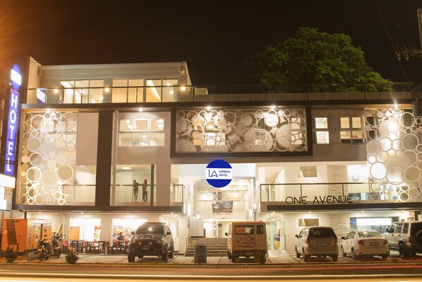A modern hotel with a prominent 'ONE AVENUE' sign, a 'HOTEL' sign, and a '1A EXPRESS HOTEL' logo. There are multiple vehicles parked in front, including cars and a van. - 1A Express Hotel (Cagayan de Oro)
