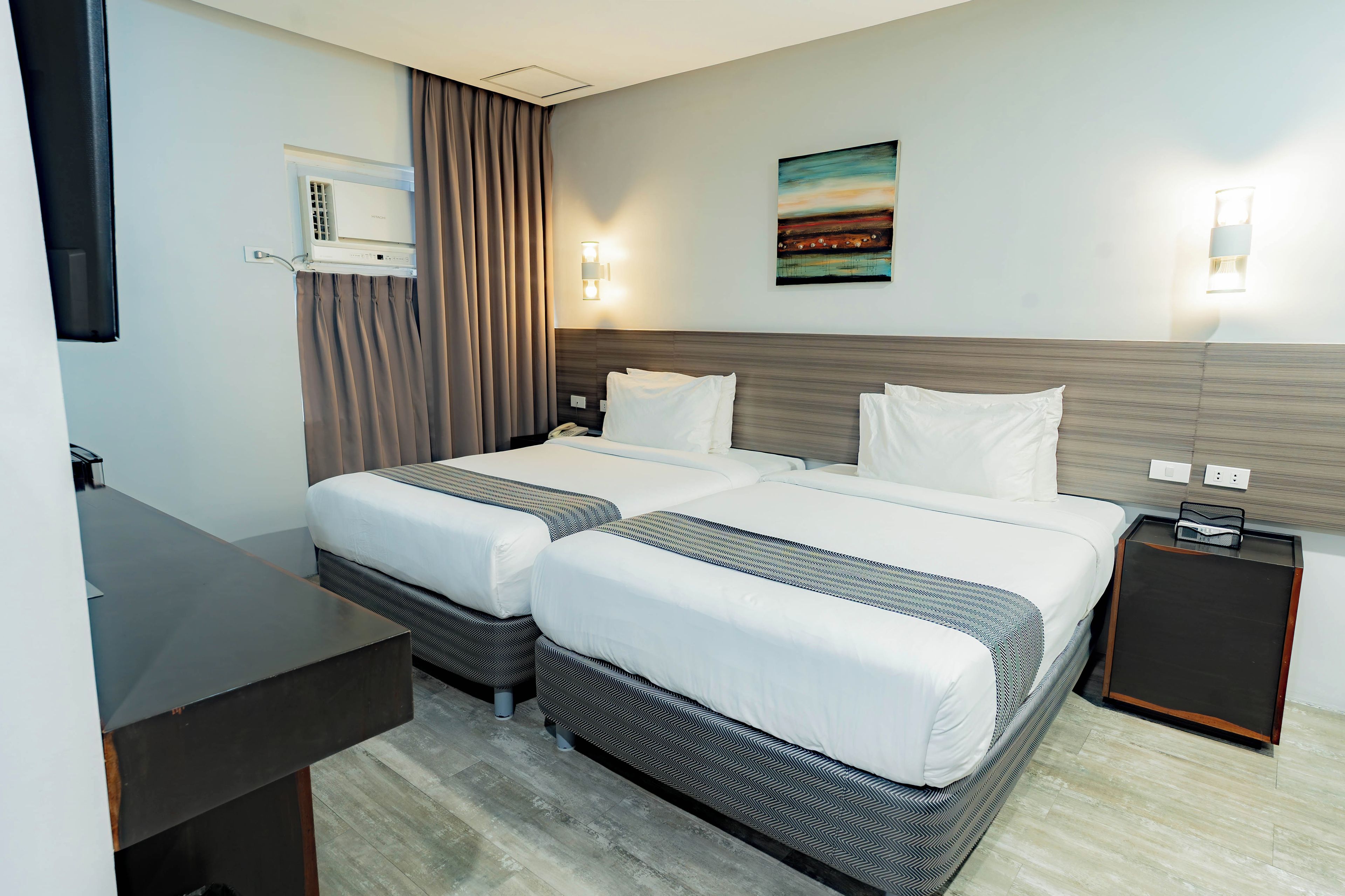 A hotel room with two beds, a desk, a nightstand, and a painting on the wall.