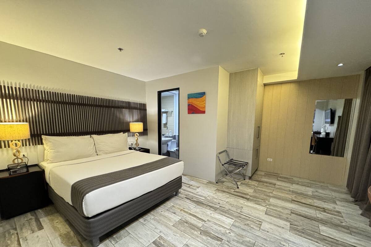 Quarto de hotel moderno com uma cama grande, mesa de cabeceira com abajur, espelho, cadeira e guarda-roupa.