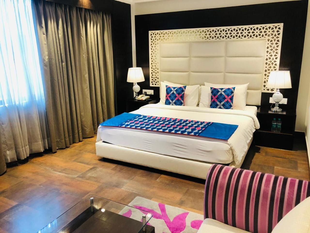 A hotel room with a large bed, a bedside table with a lamp, a striped chair, and a window with curtains.