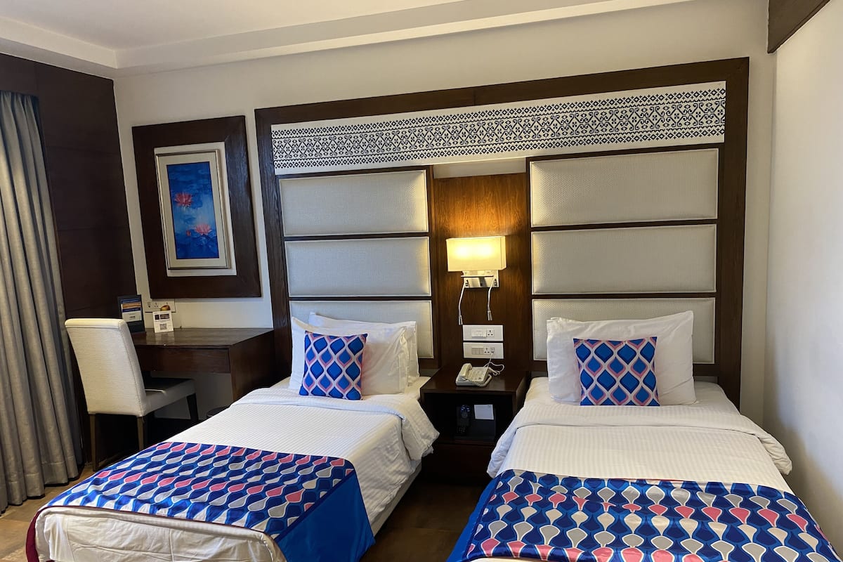 A hotel room with two beds, each with a patterned bedspread, a desk with a chair, a telephone, and a wall-mounted light.